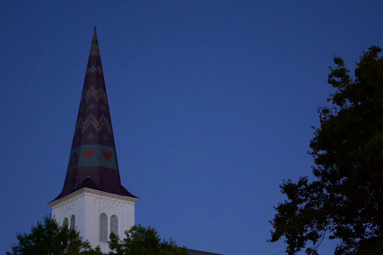 A white church steeple with a patterned, heart-studded spire against a deep blue sky at blue hour, a maple tree at the right edge